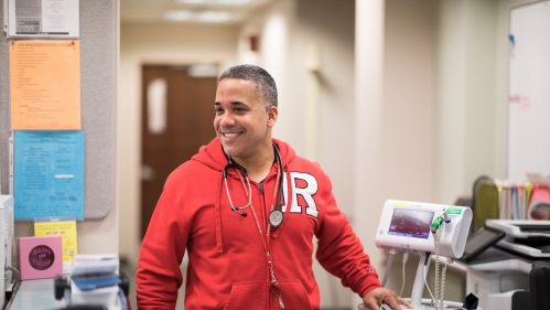 A technician wearing a red Rutgers sweatshirt wheels a monitor to a patient room in the hospital