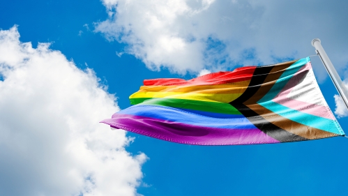 A progress pride flag waves in the wind against a blue sky with white clouds