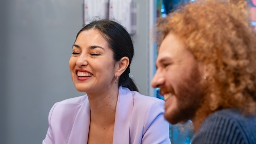 Two coworkers in an office room smile at something out of frame