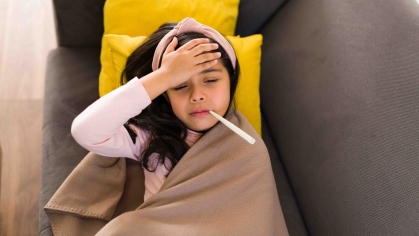 A young girl lays on a couch with her hand on her forehead and a thermometer in her mouth