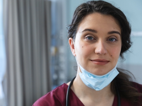 A health care worker with a mask pulled down stands in a patient room and smiles at the camera