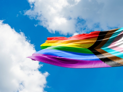 A progress pride flag waves in the wind against a blue sky with white clouds