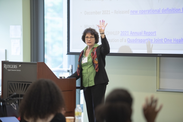 Cheryl Stroud at podium raising her left hand with large presentation screen behind her