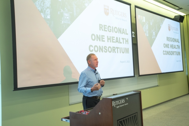 Jim Holt speaking at podium at One Health Conference with large screen in background 