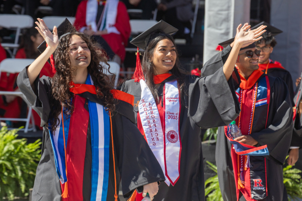Graduates in caps and gowns joyfully wave during a commencement ceremony. Two women and a man smile, exuding pride and celebration in the sunny outdoor setting.