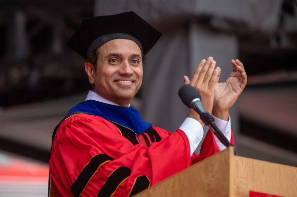 Prabhas Moghe in a red academic gown and black cap claps at a podium, smiling warmly at graduation ceremony.