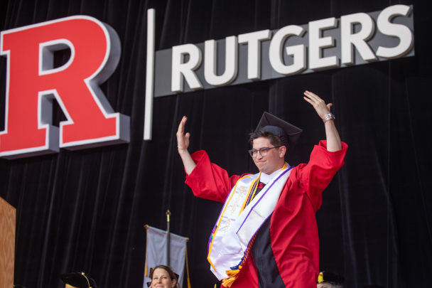 Graduating student Jack Ramirez with arms up on stage at commencement in front of a big Rutgers sign