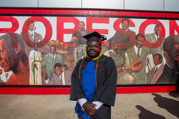 A student in commencement regalia standing in front of a mural dedicated to Paul Robeson