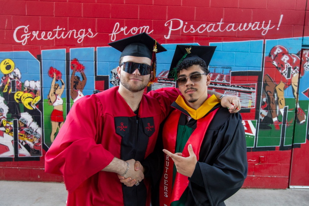 Two graduating students in regalia standing on front of a mural that says Greetings From Piscataway Rutgers
