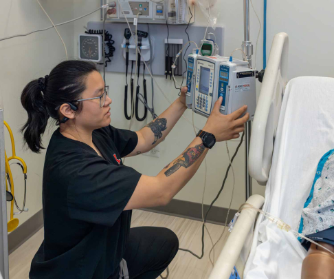 Simulation operations specialist Aj Rose programs an intravenous infusion pump, simulating medication dose and administration, next to a manikin lying in a hospital bed.