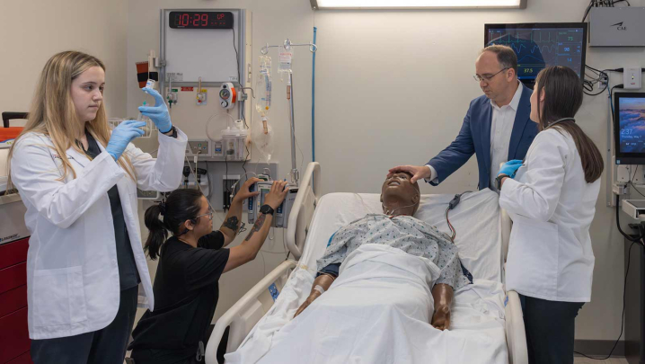 Alex Bell (left) prepares a syringe as Aj Rose programs an intravenous infusion pump while Les Barta and Amanda Sallemi stand over a manikin lying in a hospital bed.